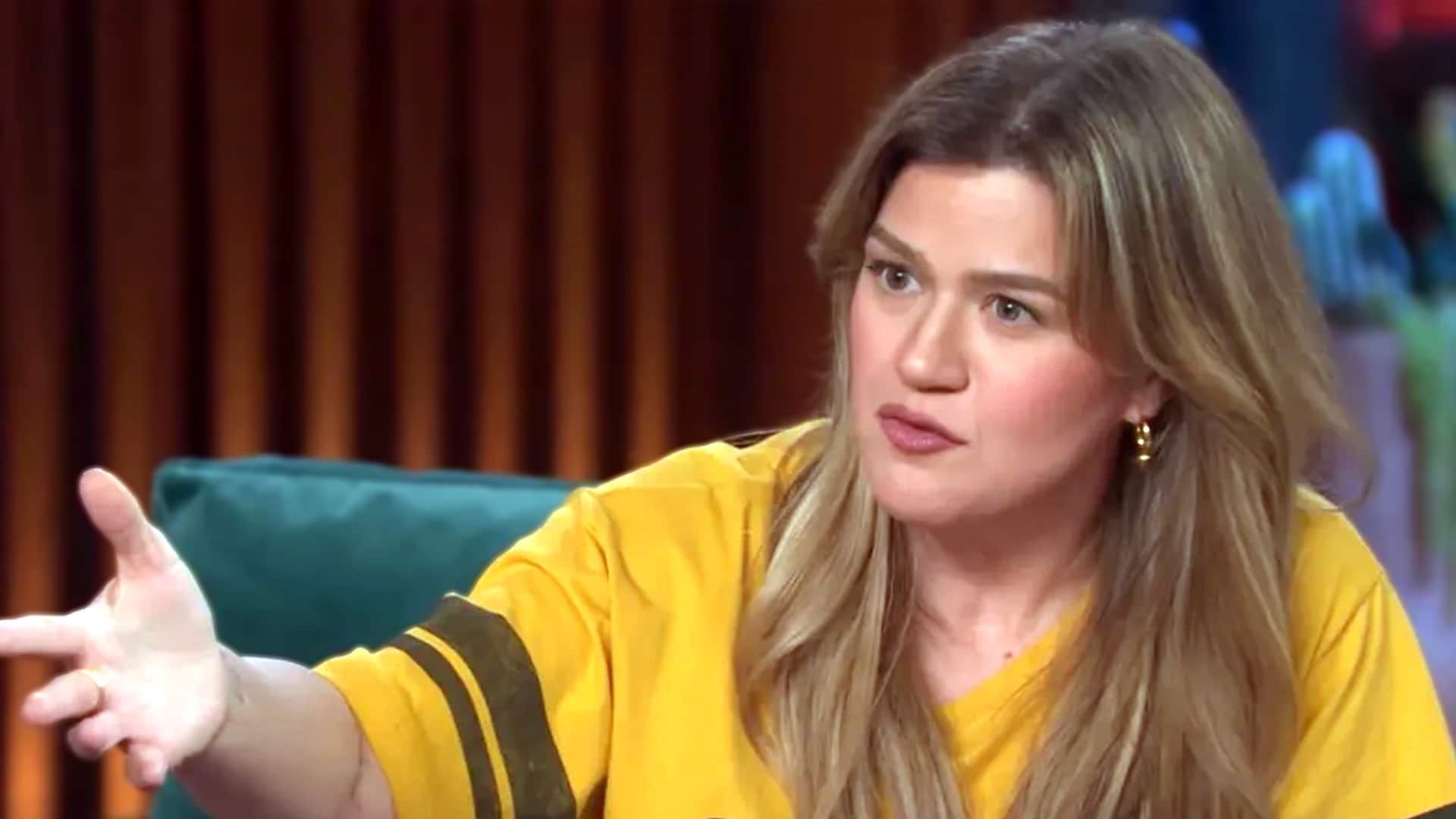 Kelly Clarkson gestures emphatically during an interview, revealing she forfeited songwriting royalties worth hundreds of thousands (or millions) on "My Life Would Suck Without You" rather than share official credit with producer Dr. Luke.