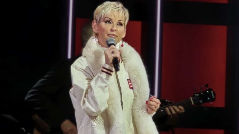Lorrie Morgan delivering a breathtaking performance of "Something in Red" at the Grand Ole Opry 100th anniversary celebration on November 28, 2025, wearing a white fur-trimmed outfit while holding a microphone on the historic Nashville stage.