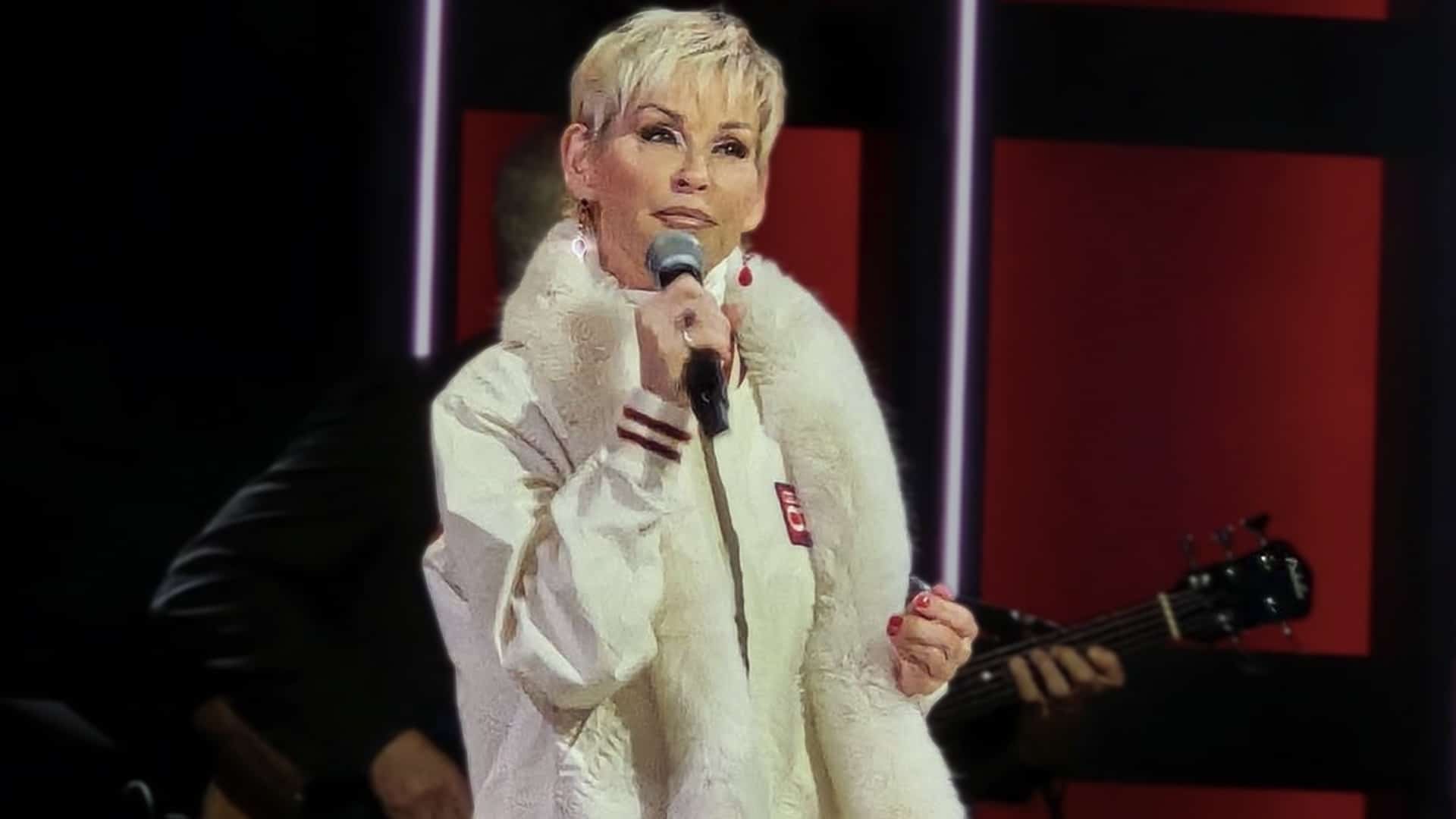 Lorrie Morgan delivering a breathtaking performance of "Something in Red" at the Grand Ole Opry 100th anniversary celebration on November 28, 2025, wearing a white fur-trimmed outfit while holding a microphone on the historic Nashville stage.