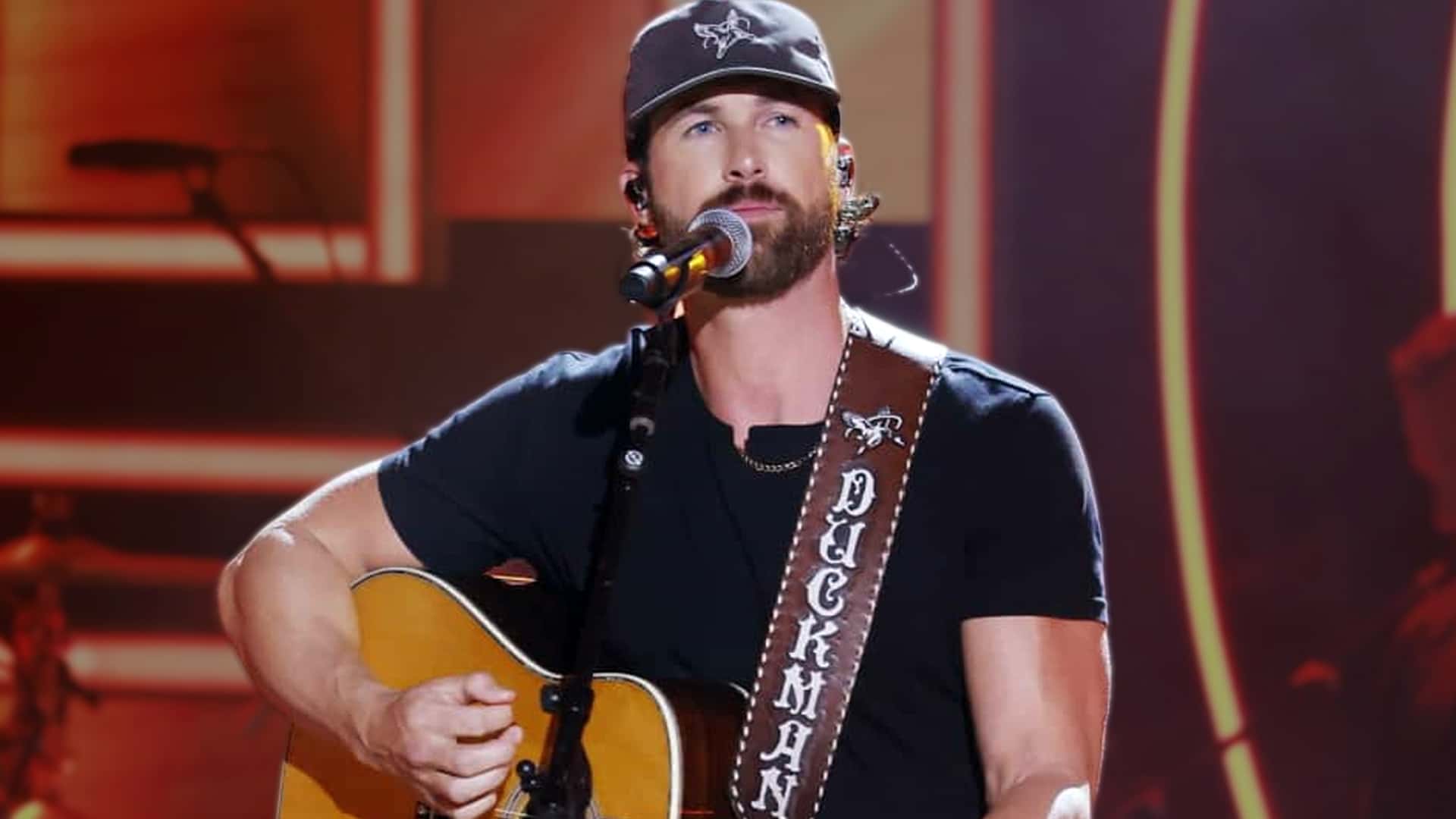 Riley Green performs live on stage in black cap and t-shirt with "Deckhand" guitar strap, becoming the first artist since Taylor Swift to score back-to-back solo-written No. 1s on Billboard Country Airplay with "Don't Mind If I Do" in 2025.