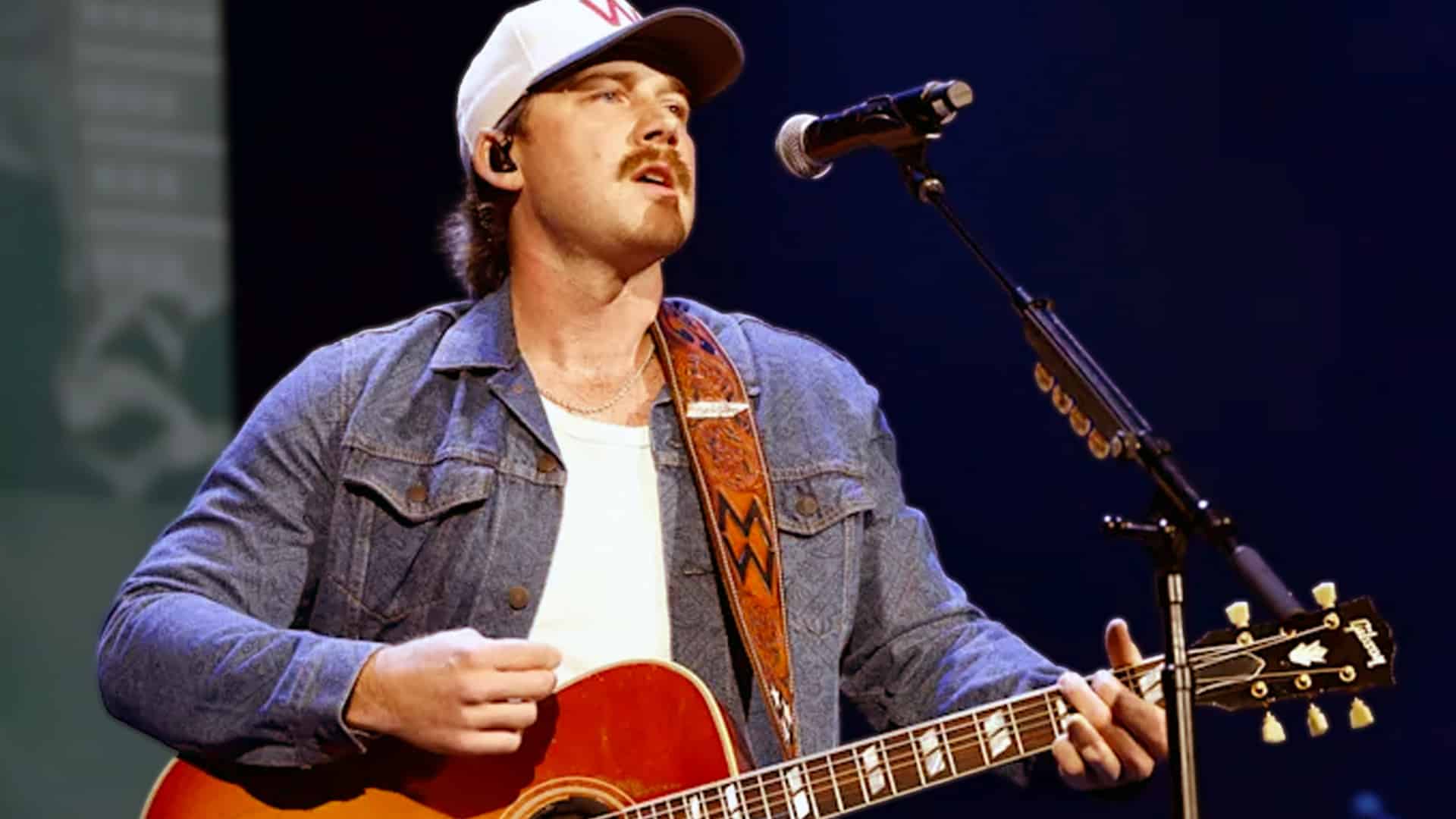 Morgan Wallen performing live on stage in a white W cap, denim jacket, and mustache, passionately singing into a microphone while strumming a red Gibson acoustic guitar, celebrating his album "I'm The Problem" as Spotify's #1 most-streamed U.S. album of 2025, outpacing Taylor Swift and Kendrick Lamar.