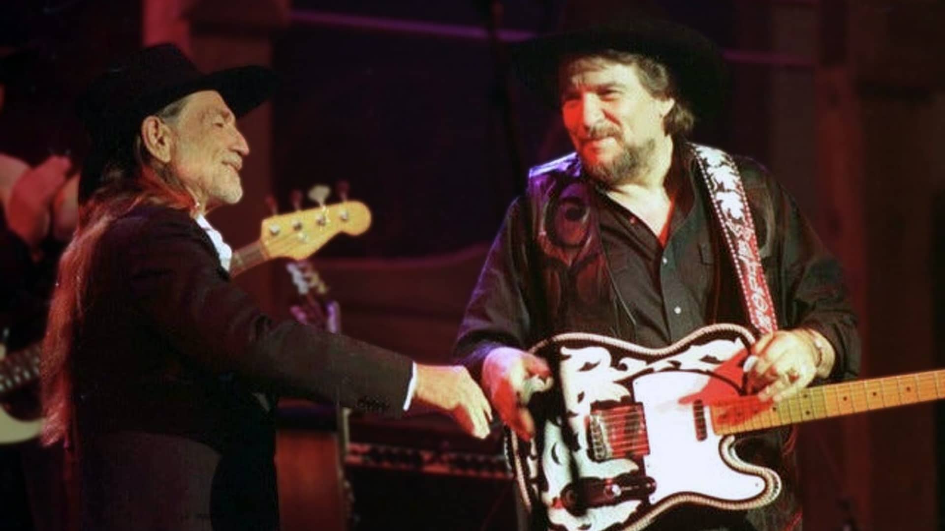 Waylon Jennings and Willie Nelson share a moment on stage, with Waylon in black hat and suit shaking hands with Willie holding his signature Trigger guitar, symbolizing the outlaw country bond when Waylon fought Columbia Records executives in 1975 to release Willie’s raw Red Headed Stranger album untouched.