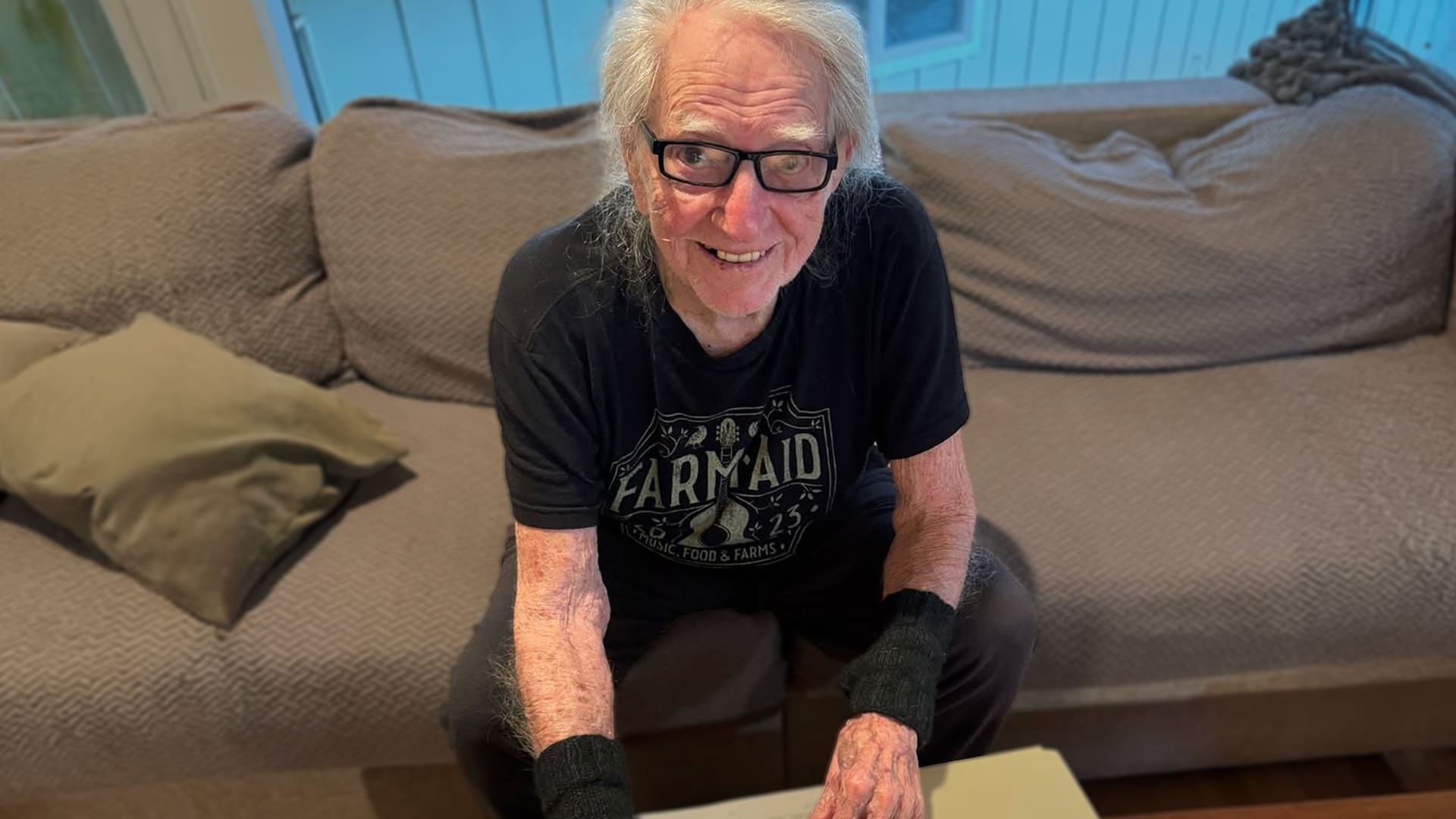 Willie Nelson at 92, smiling in a Farm Aid 2023 t-shirt and wrist warmers while sitting on a couch, as he helps distribute $1.3 million in 2025 Farm Aid grants to support family farms and rural America.