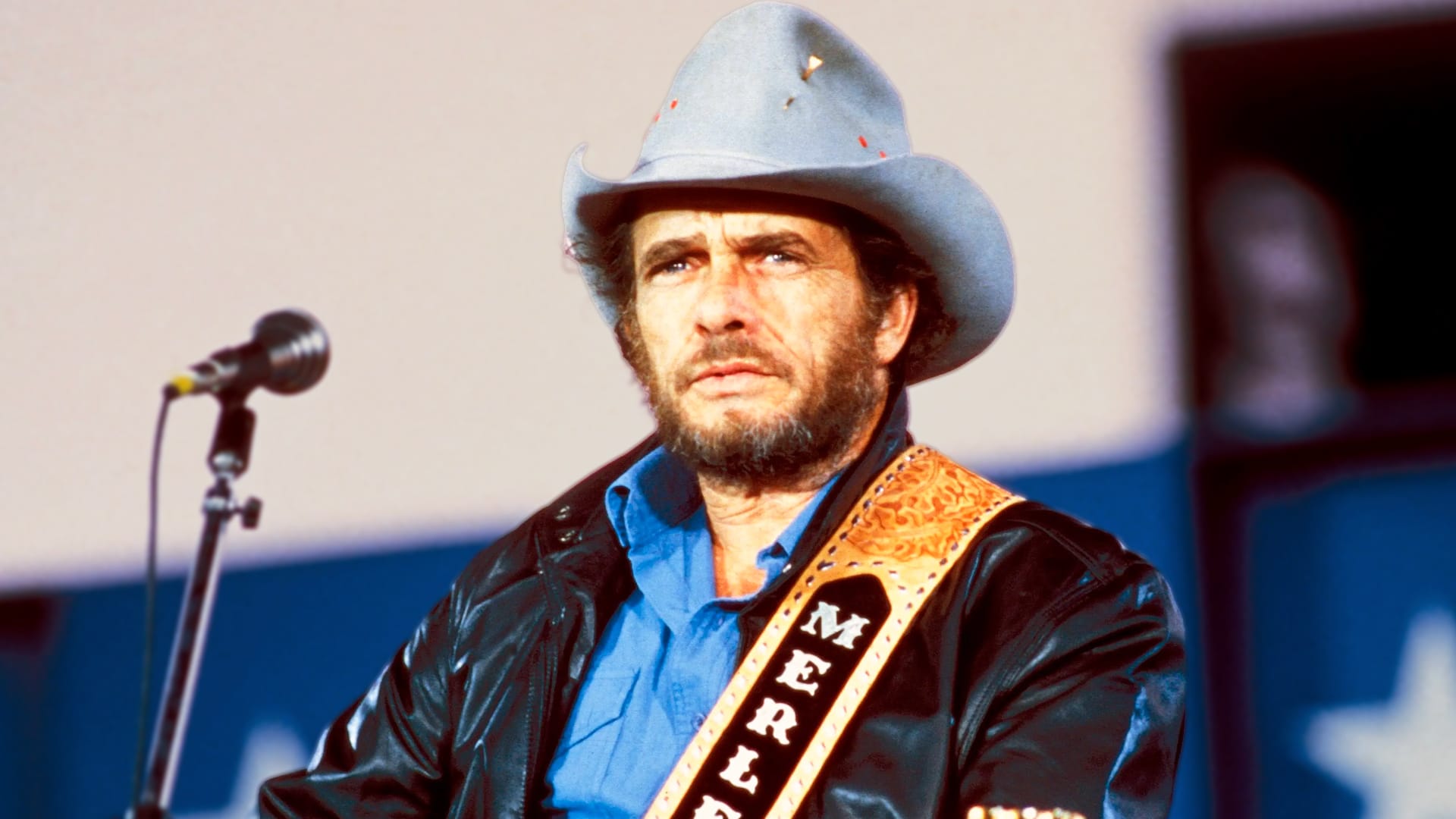 Merle Haggard performs "If We Make It Through December" on stage in light cowboy hat and leather jacket with "Merle" guitar strap, the classic 1973 song often mistaken for Christmas music but actually a raw working-class story of survival and heartbreak.