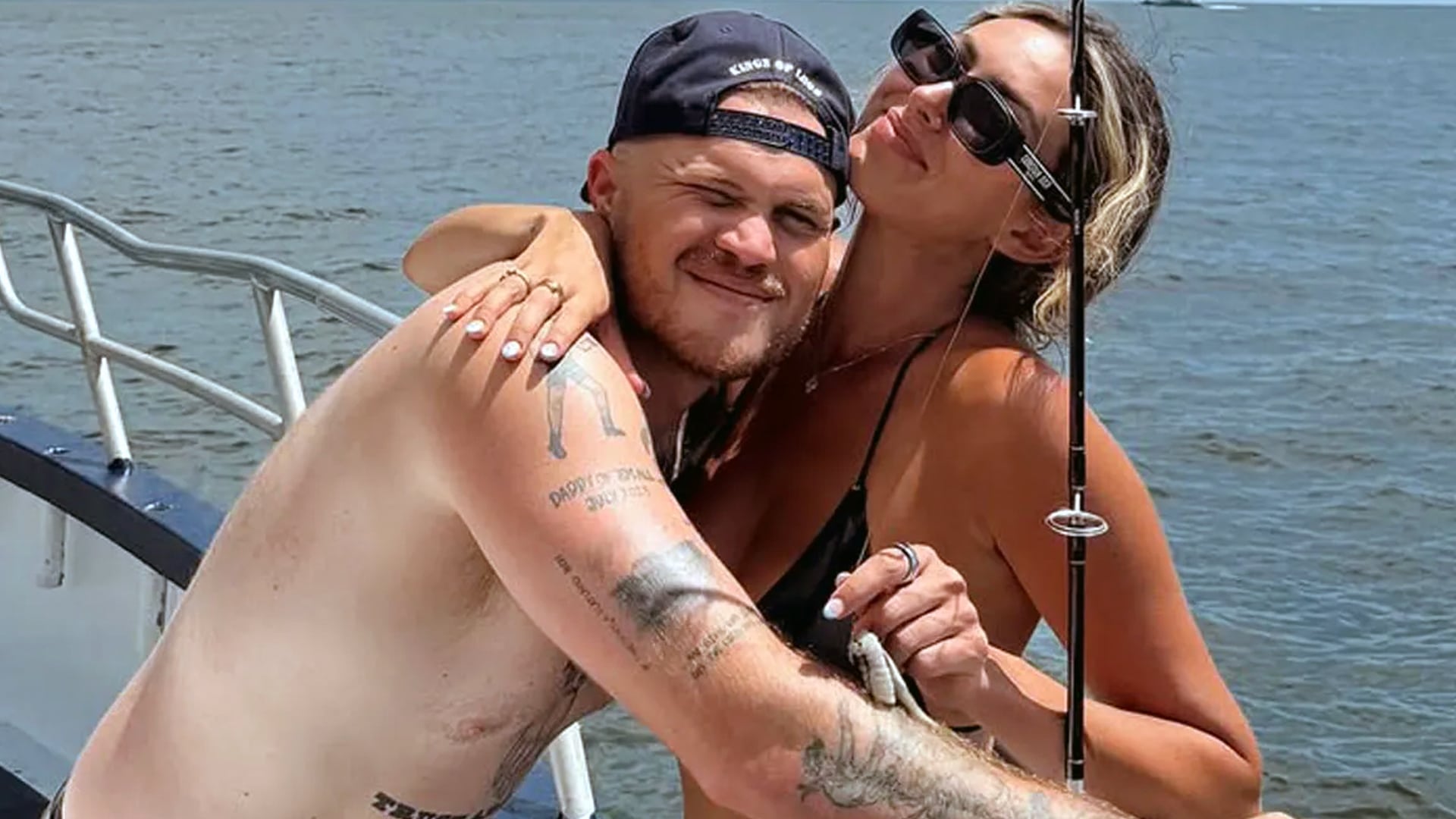 Zach Bryan and new wife Samantha Leonard share a joyful moment on their wedding day in Spain, Samantha kissing his cheek on a boat as Zach wears a "Cases of Love" cap, following reports of their secret 2025 ceremony in San Sebastián.