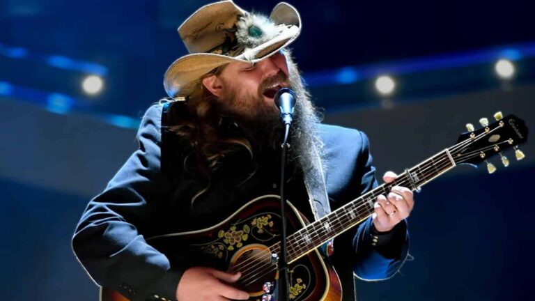 Chris Stapleton performs "Tennessee Whiskey" on stage in cowboy hat and dark jacket with guitar, as the song becomes the first country track to earn RIAA double diamond status with over 20 million units sold in 2026.