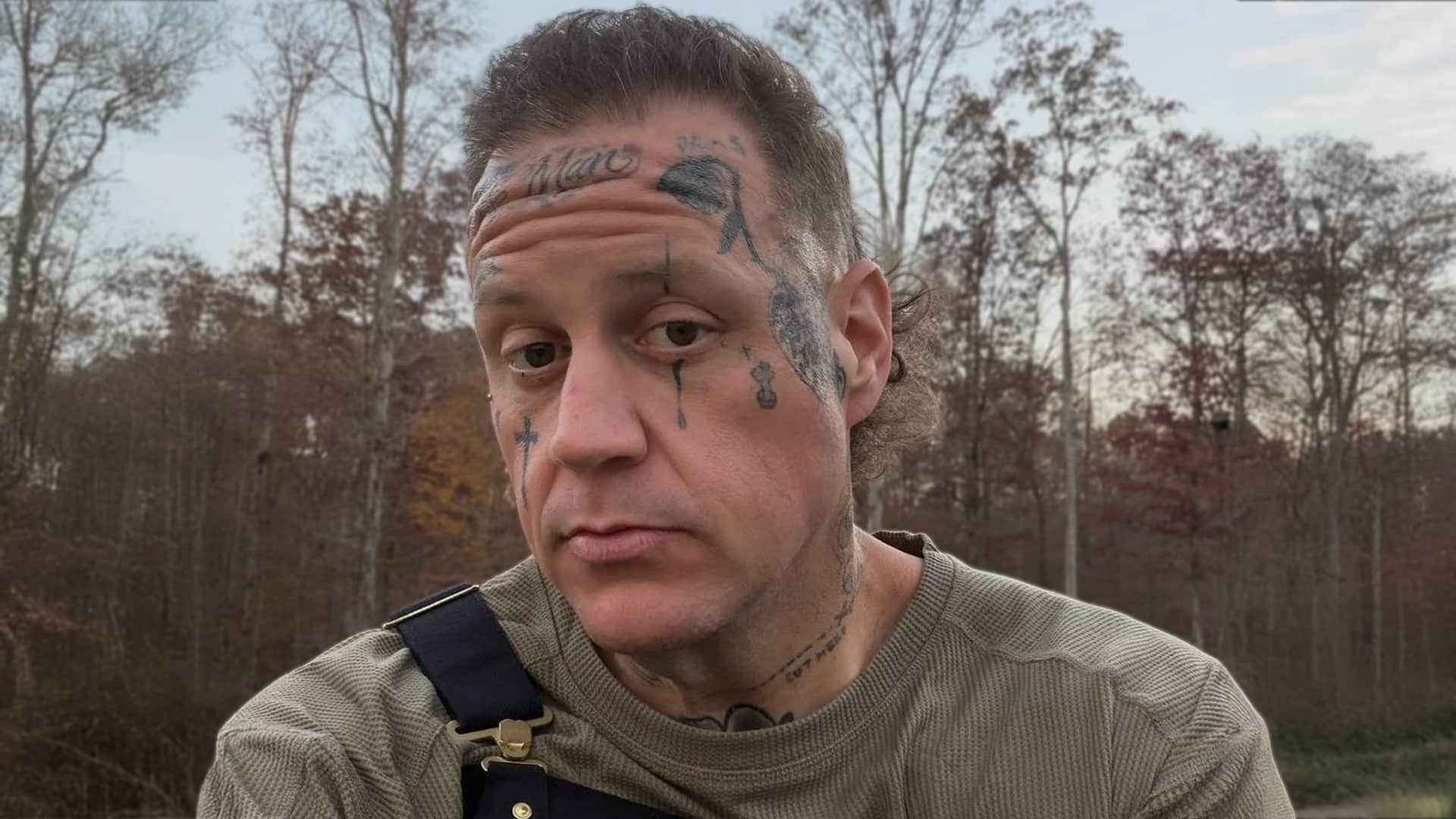 Jelly Roll with extensive face and neck tattoos in gray sweater and overalls outdoors, revealing in 2026 Men's Health interview he has considered tattoo removal not from regret but as part of his personal growth and life transformation.