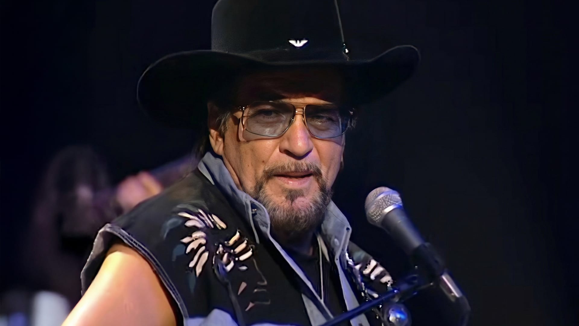 Waylon Jennings performs from a seated spot, still commanding the stage with outlaw grit that defined his final chapter in 2000.