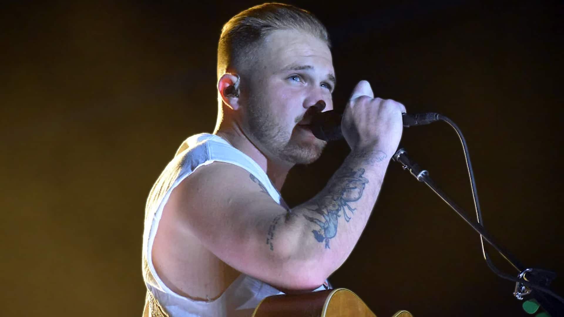 Zach Bryan performs intensely on stage in white tank top with guitar, as his new 2026 track "Skin" from With Heaven on Top appears to be a sharp lyrical goodbye to ex Brianna Chickenfry, referencing tattoos and past pain.