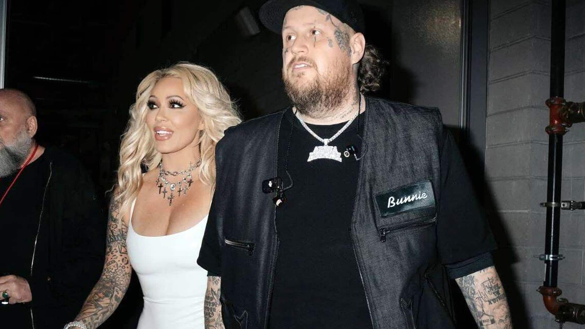 Bunnie XO and Jelly Roll walk arm-in-arm at event, Bunnie in white dress and bold makeup, Jelly Roll in black vest with "Bunny" patch, tattoos visible, as they embrace faith as "baby Christians" after rediscovering God.