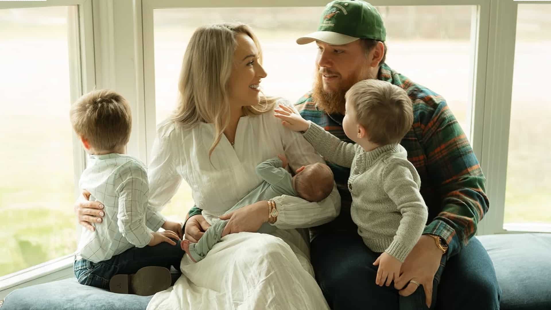 Luke Combs and wife Nicole smile with newborn son Chet Wiley Combs in cozy family portrait, big brothers Tex and Beau gazing at baby brother on window seat, celebrating arrival of third boy announced February 19, 2026.