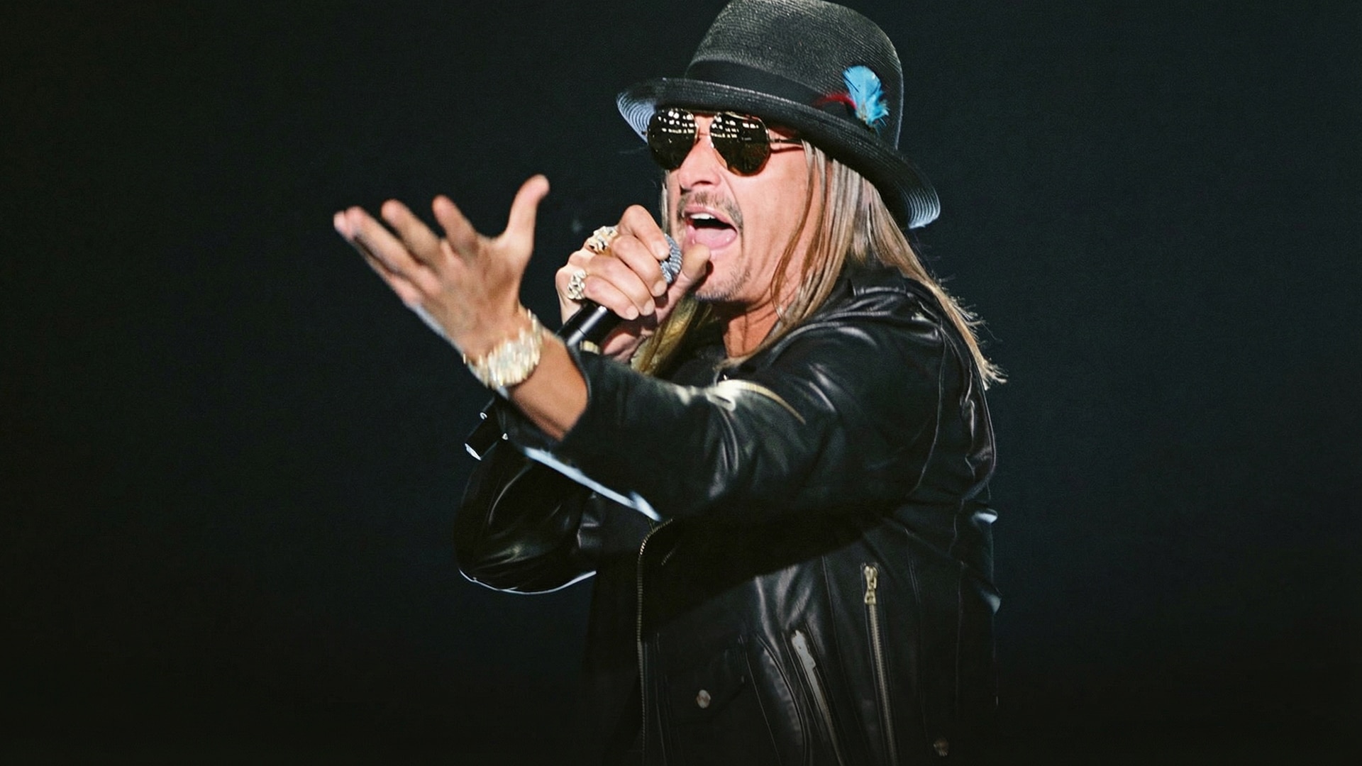 Kid Rock performs energetically on stage in black leather jacket, feathered black hat, aviator sunglasses, and rings, microphone in hand during Freedom 250 Tour, defending $5,000 front-row ticket prices for premium proximity.