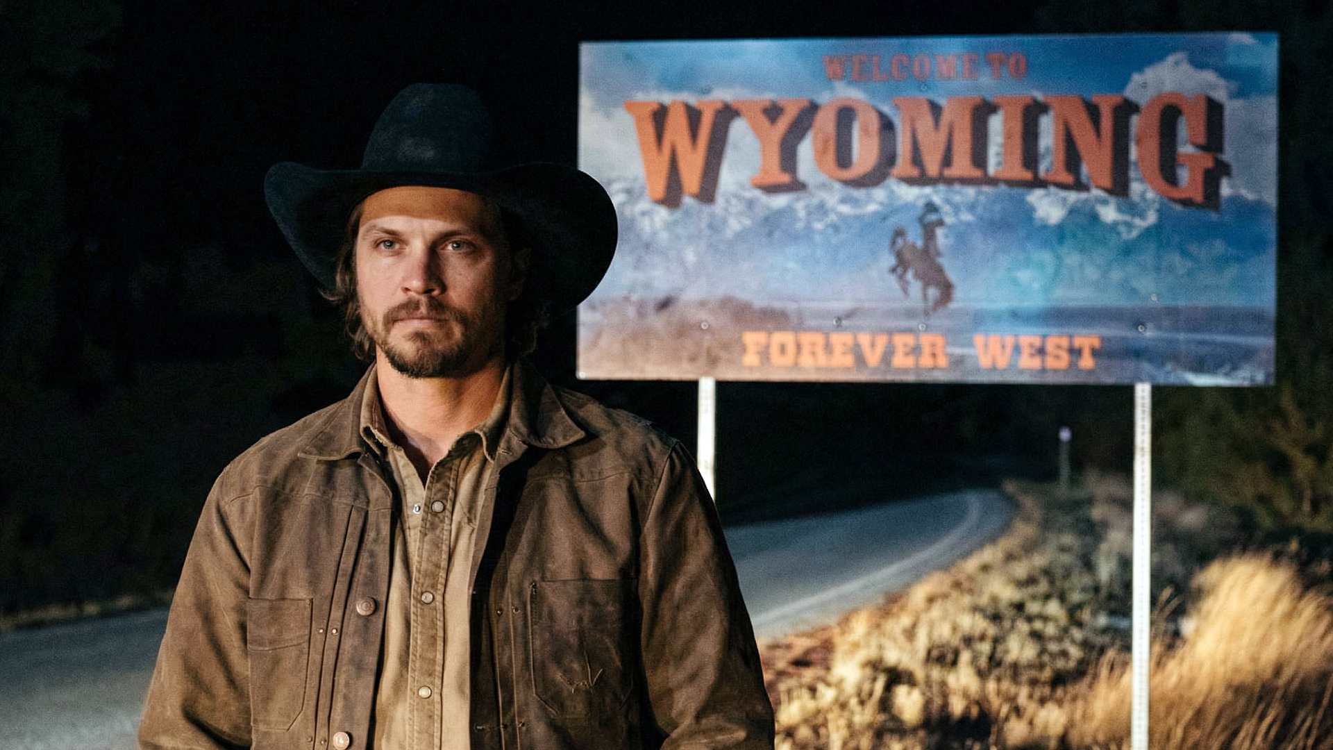 Luke Grimes as Kayce Dutton stands before Wyoming welcome sign at night in Marshals Yellowstone spinoff, cowboy hat and jacket, revisiting the haunting train station legacy in episode two "Zone of Death."