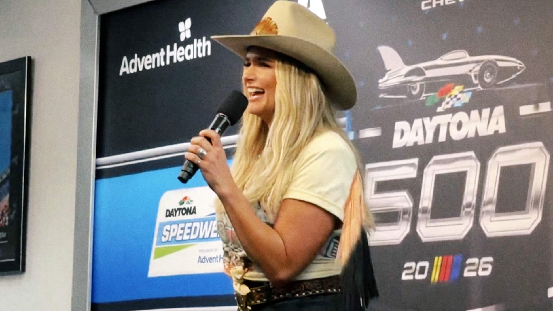Miranda Lambert speaks enthusiastically at a press conference podium in front of a Daytona 500 banner, wearing a cowboy hat and fringed outfit, ready to kick off the 2026 NASCAR season with her pre-race performance.
