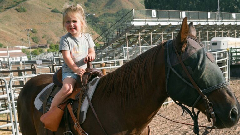Young Oaklynn Rae Domer, 3-year-old daughter of WPRA champion Kelsie Domer, smiles atop brown horse in saddle at rodeo arena with hills behind, days before tragic February 19, 2026 accident sparks widespread pink tributes in mourning.