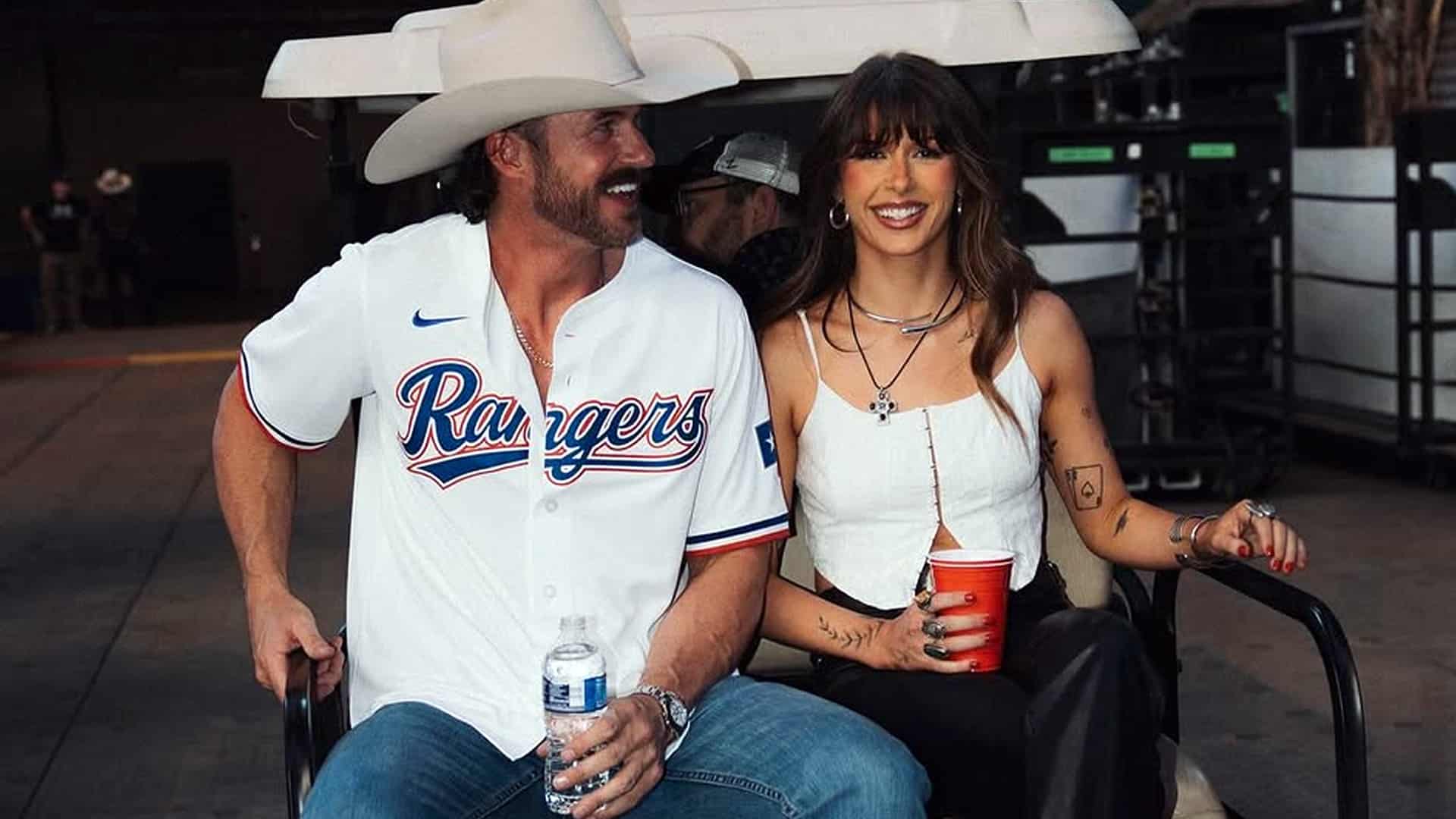 Riley Green and Ella Langley smile together in golf cart at event, Riley in white Texas Rangers jersey and cowboy hat, Ella in white top holding red cup, celebrating her "Choosin' Texas" No. 1 chart success and Alabama roots.