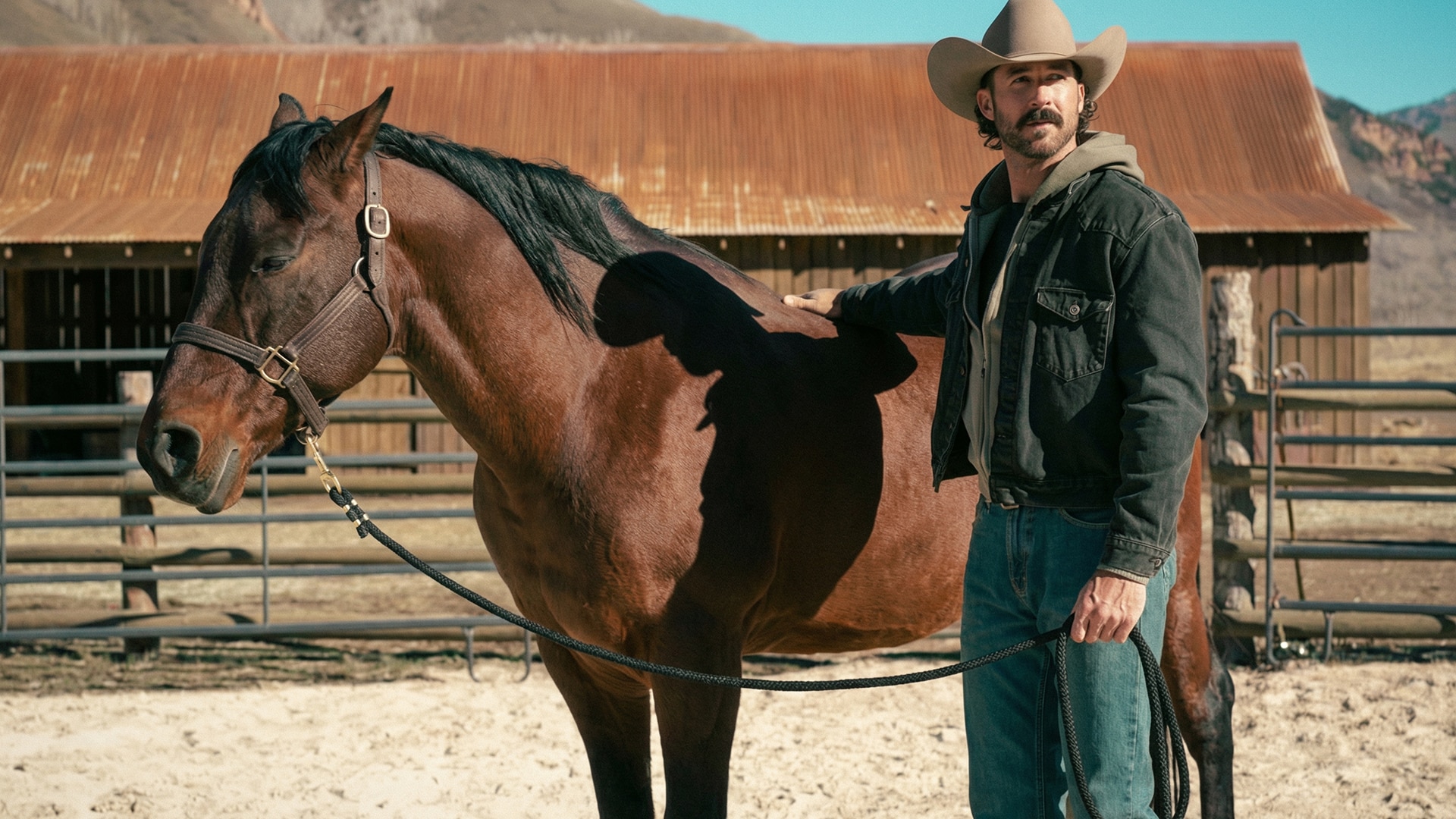 Riley Green makes acting debut as former Navy SEAL Garrett in Yellowstone spinoff Marshals, standing with brown horse by rustic barn in cowboy hat, denim jacket, and mustache in official promo image ahead of March 1 premiere.