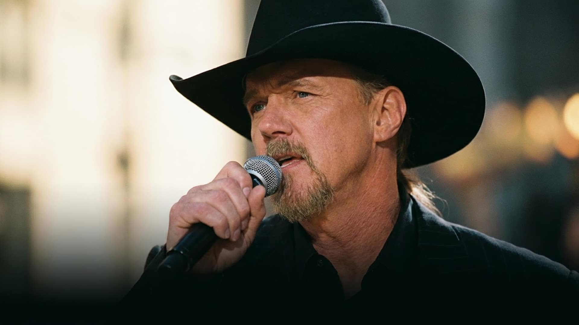 Trace Adkins performs live on stage in black pinstripe suit and cowboy hat, microphone in hand under colorful lights, hinting 2026 30th Anniversary Tour may be his final road run at age 64.